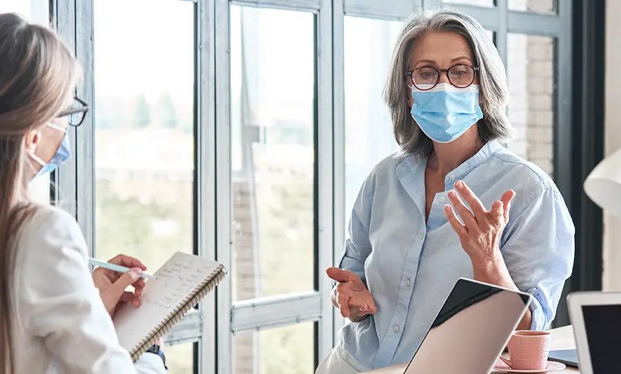 Older female talks to a nurse while both wear masks